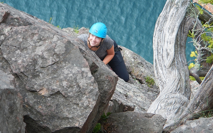 A person wearing safety gear is secured by ropes as they look up at the camera and smile while rock climbing. They are high above a blue body of water. 